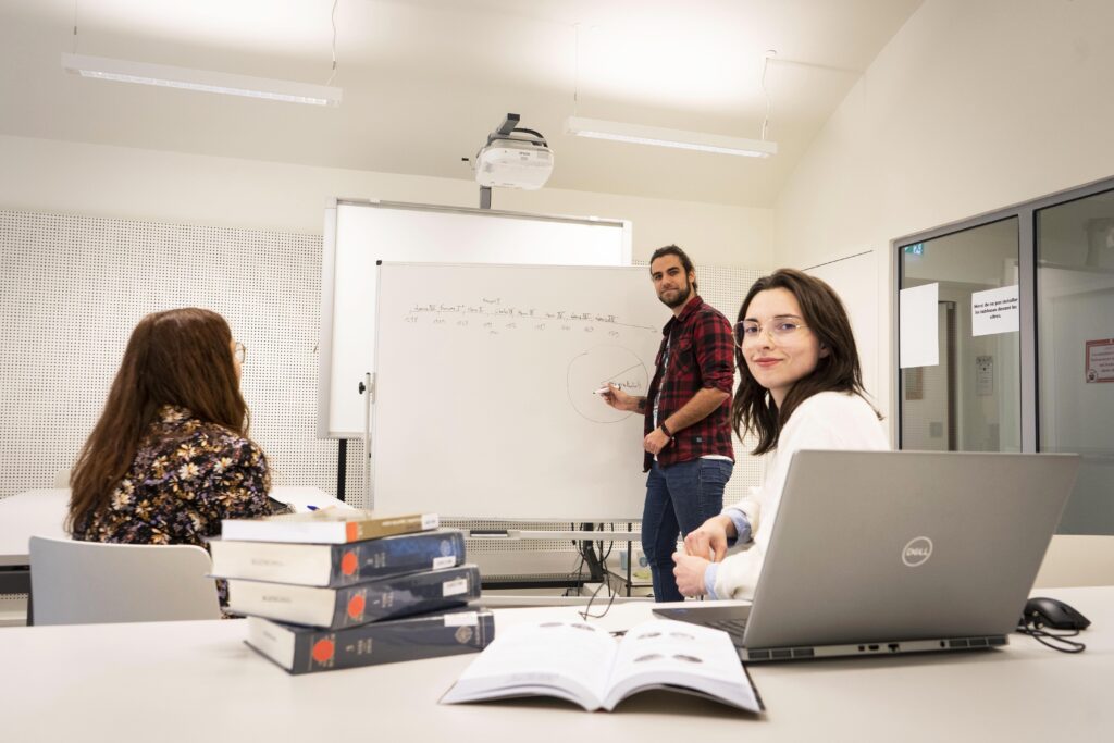 photo à l'intérieur d'une salle de travail en groupe. une personne écrit au tableau blanc, une autre, de dos, regarde le tableau et une troisième est devant un ordinateur portable